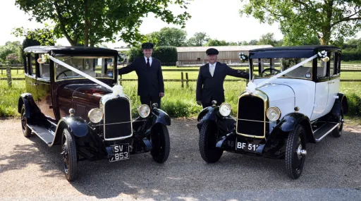 Two Vintage Wedding Cars with chauffeur standing by the vehicles. Both decorated with matching pair of white ribbons.